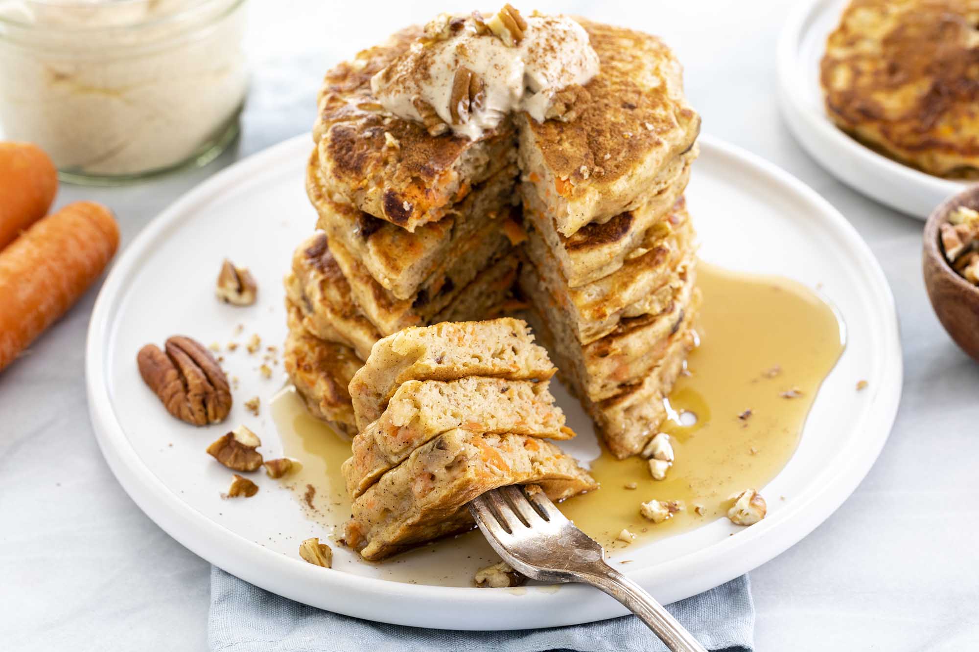 Tall stack of buttermilk carrot cake pancakes with a cream cheese topping, chopped pecans and a puddle of syrup at the bottom of the plate. Whole carrots, a glass of cream cheese topping, more pancakes and a bowl of chopped pecans surround the plate.