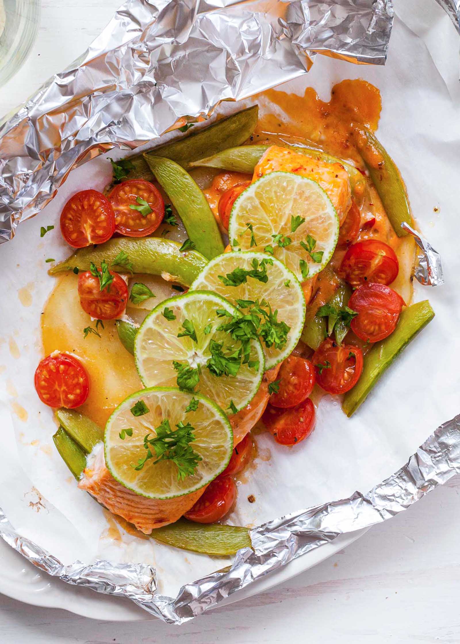 Top view of the best healthy salmon. A fillet is in the middle of an open foil packet with grape tomatoes, sugar snap peas, and sauce around it. Thinly sliced limes and chopped cilantro are on top of the salmon.