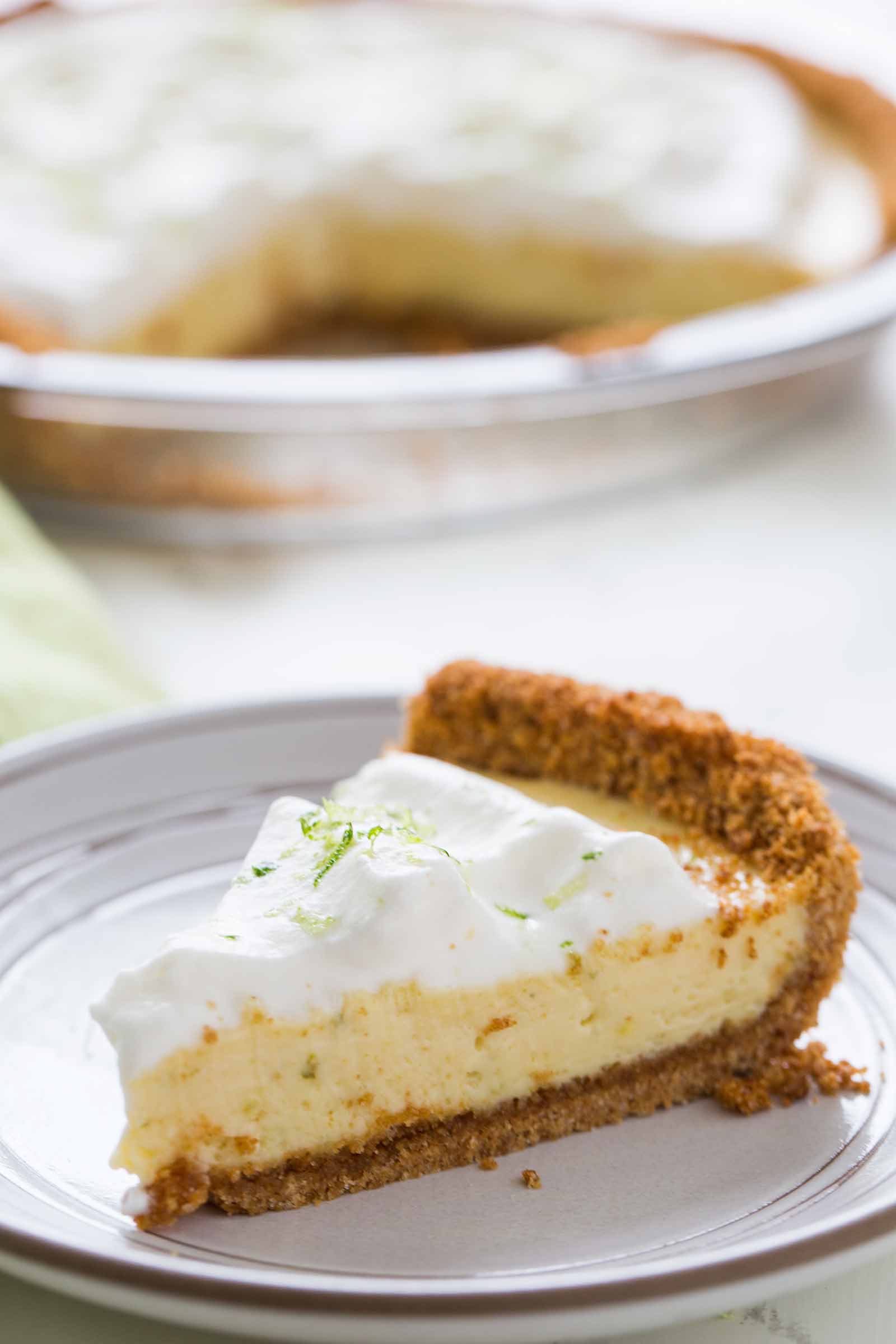 One slice of the best key lime pie on a plate with the rest of the pie in the background