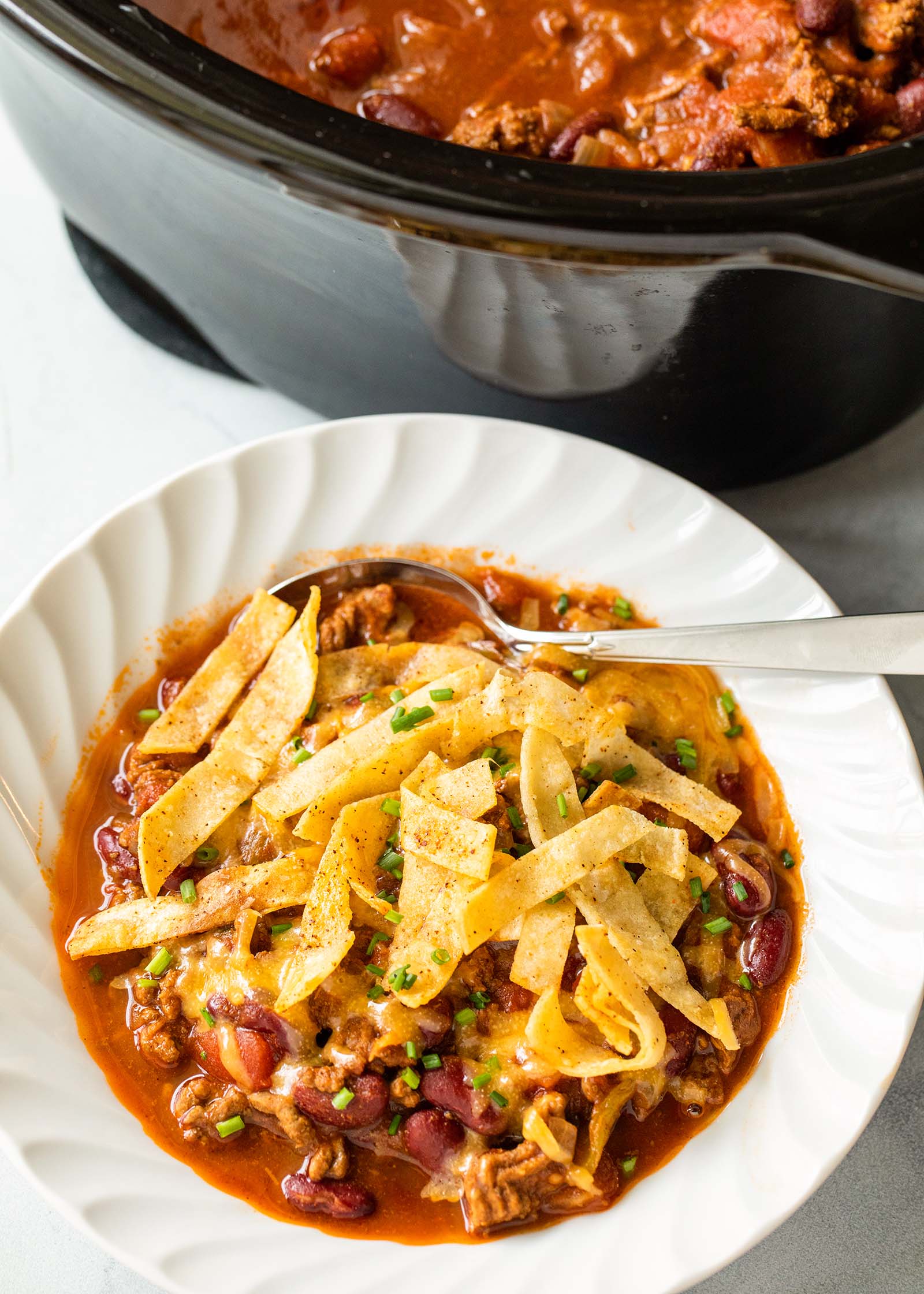 A bowl of Tailgate Beef Chili Recipe topped with fried tortilla strips with a crock pot set above it.