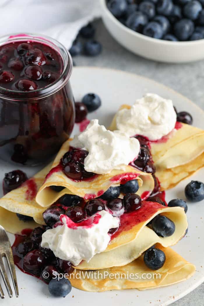 Blueberry Crepes on a white plate with whipped cream and blueberries surrounding it