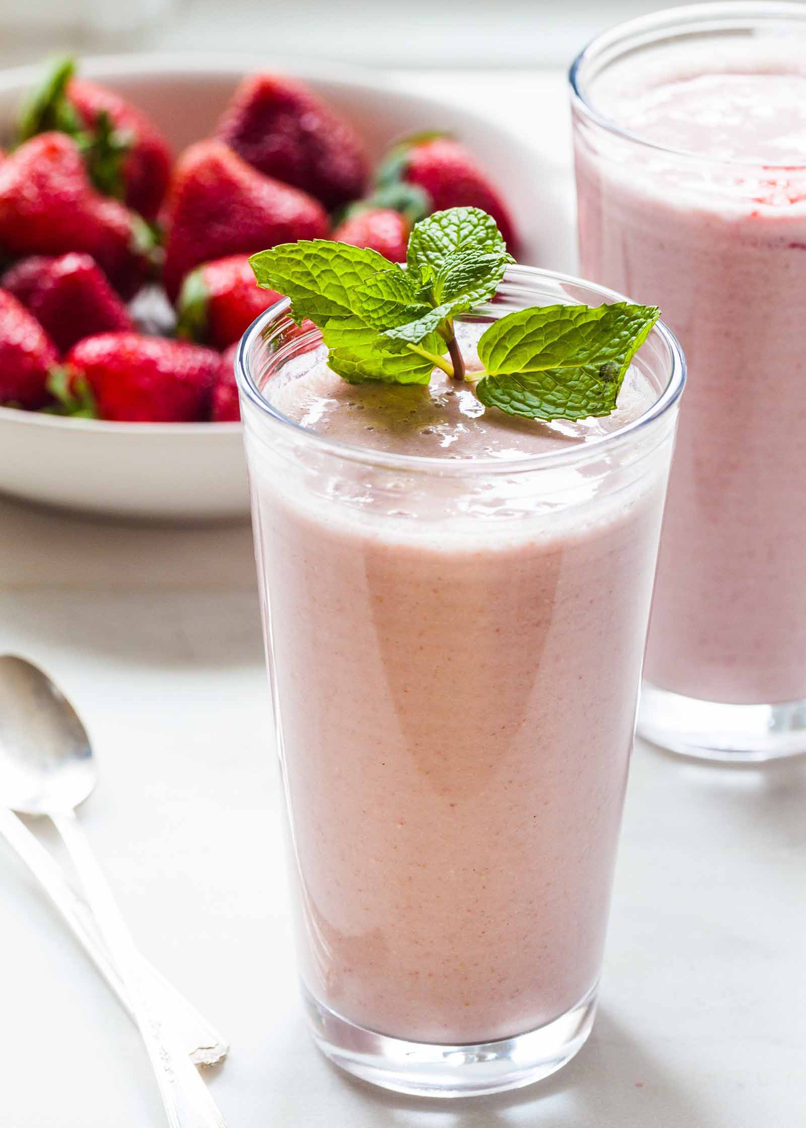 Vertical view of two best strawberry almond oat smoothies. A sprig of mint sits on the top of one glass and the other glass is in partial view to the right. Behind the first glass is a bowl of strawberries. Two spoons are in partial view to the left of the glass.