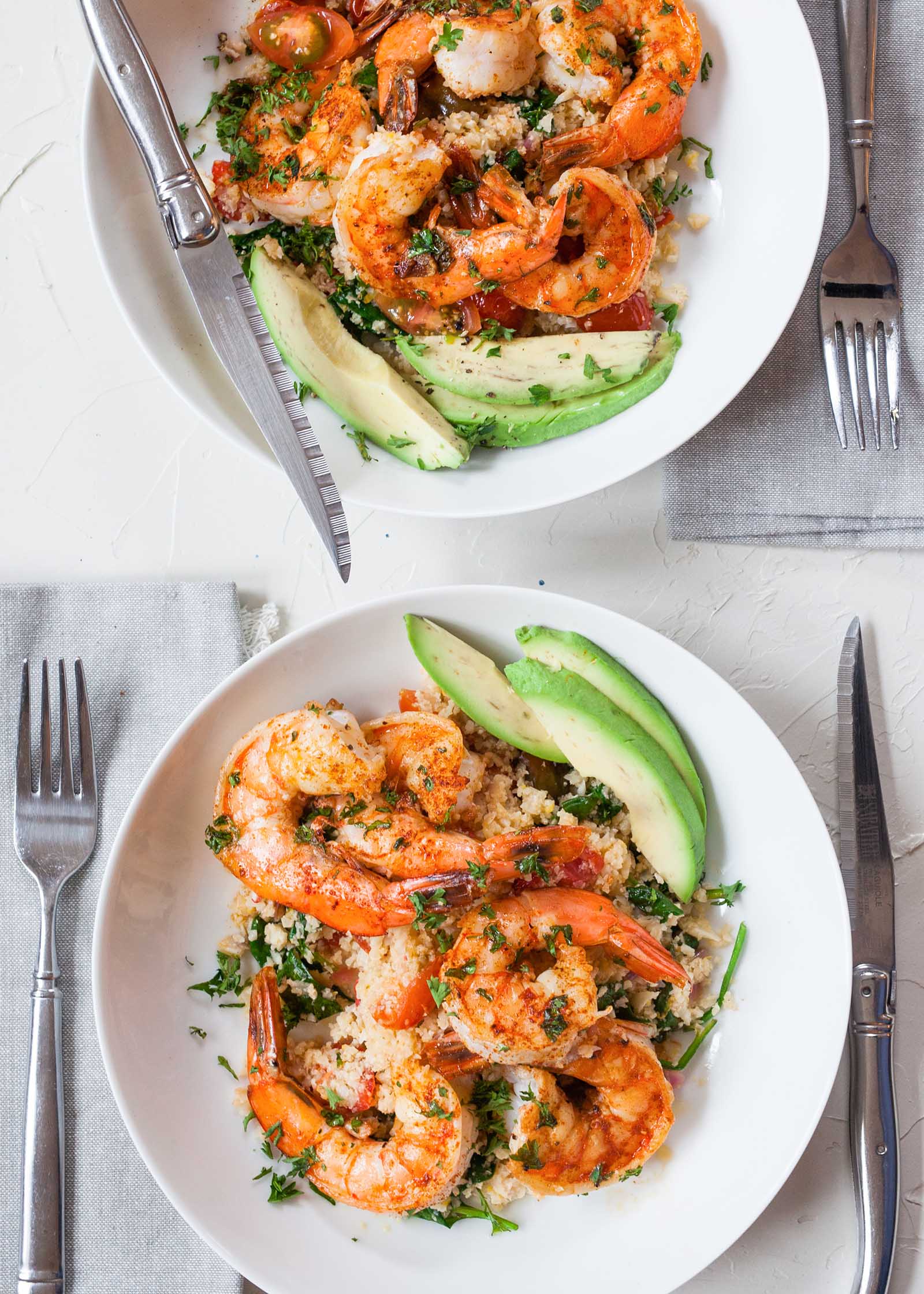 Vertical photo of two table settings of garlic shrimp cauliflower rice bowls. Avocado slices and herbs are in the bowls as well. A fork, knife and napkin are at each place setting.
