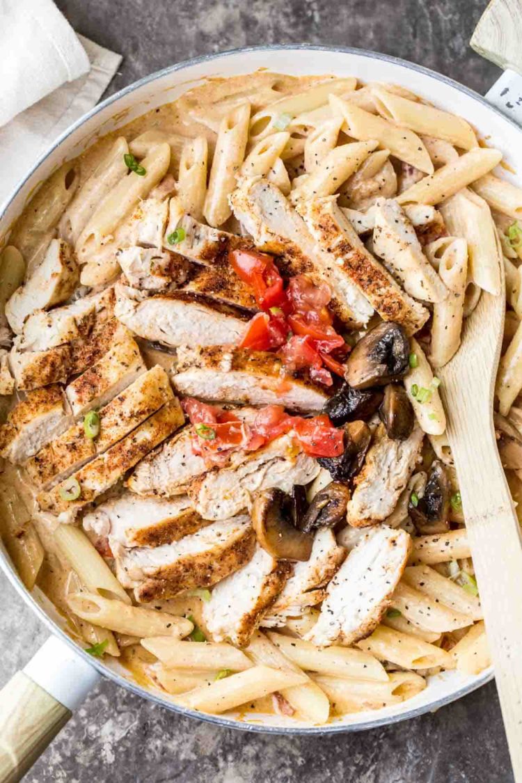 Receta de pasta cremosa de pollo cajún en una sartén cubierta con pollo, champiñones, tomates y una cuchara de madera.