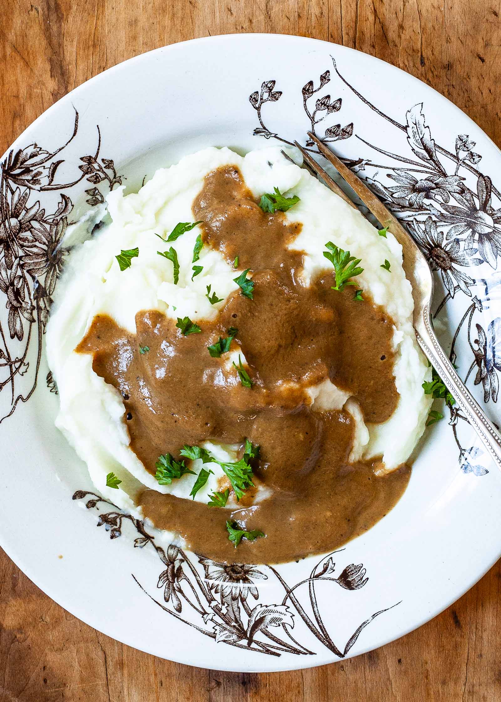 Easy Vegan mushroom gravy poured over a pile of mashed potatoes on a white and brown plate with a fork.