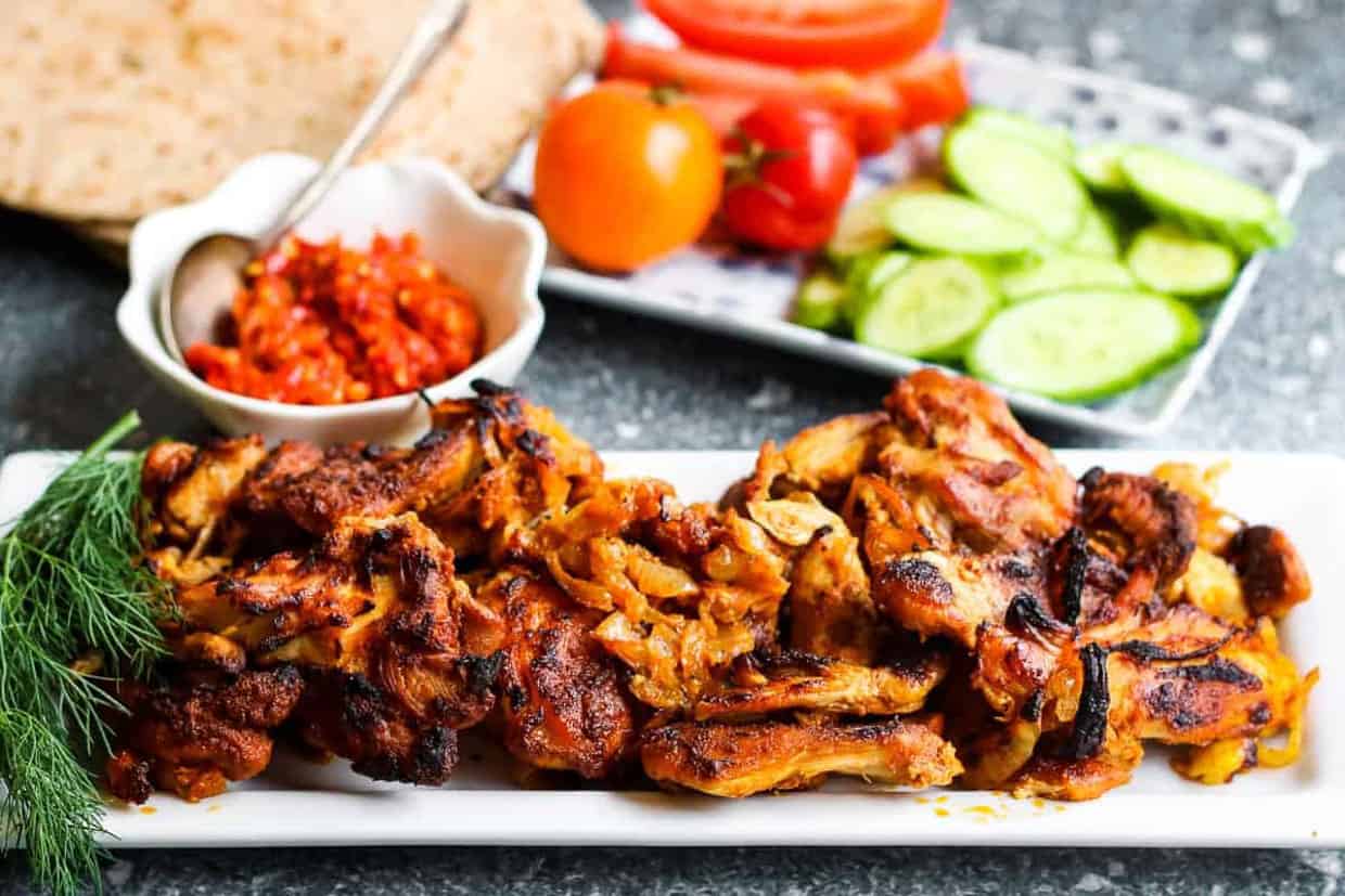 low angle shot of chicken shawarma on a white platter with harissa paste and tomatoes and cucumbers in the background.